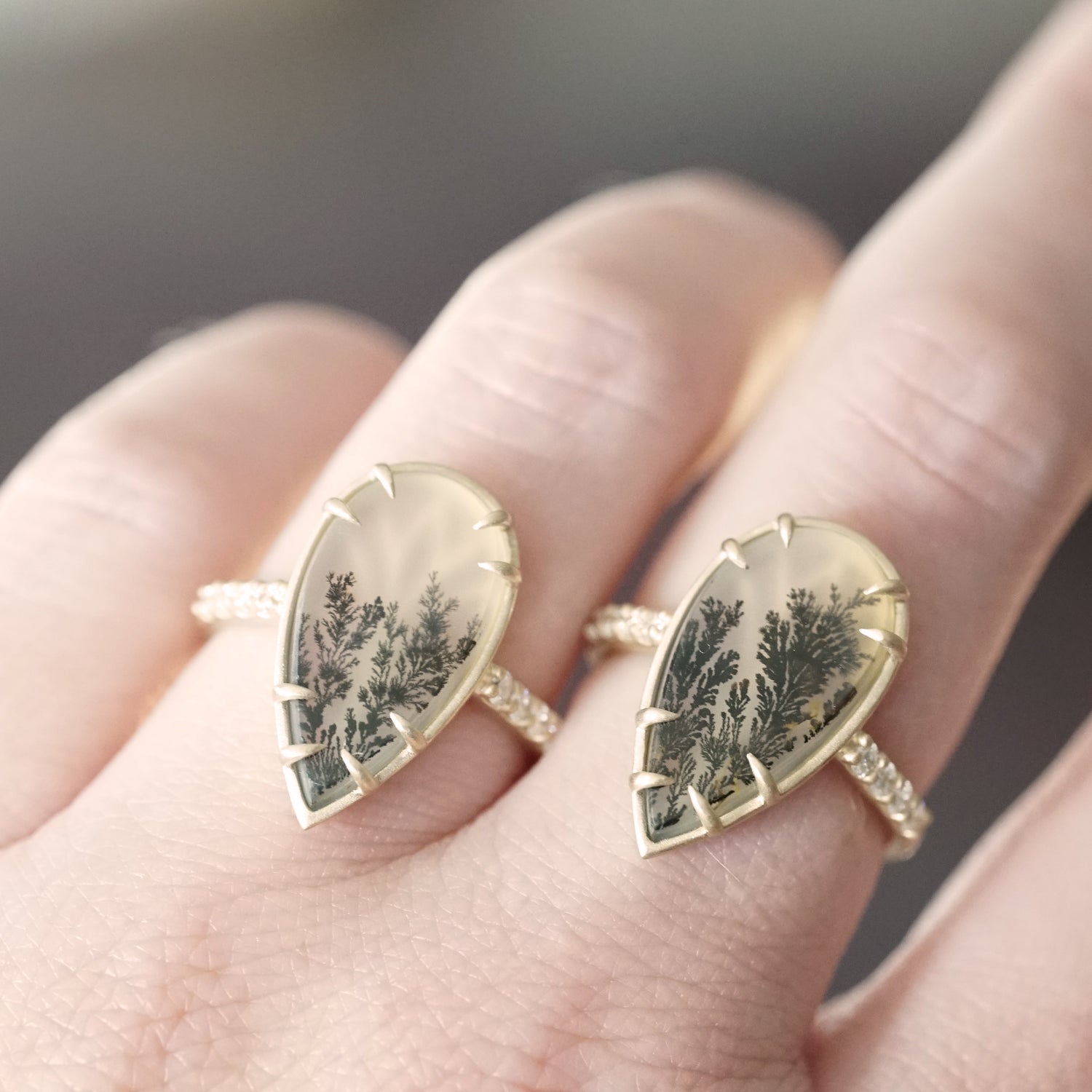 two yellow gold rings with dendritic agate center stones and diamond shanks on a light skinned left hand