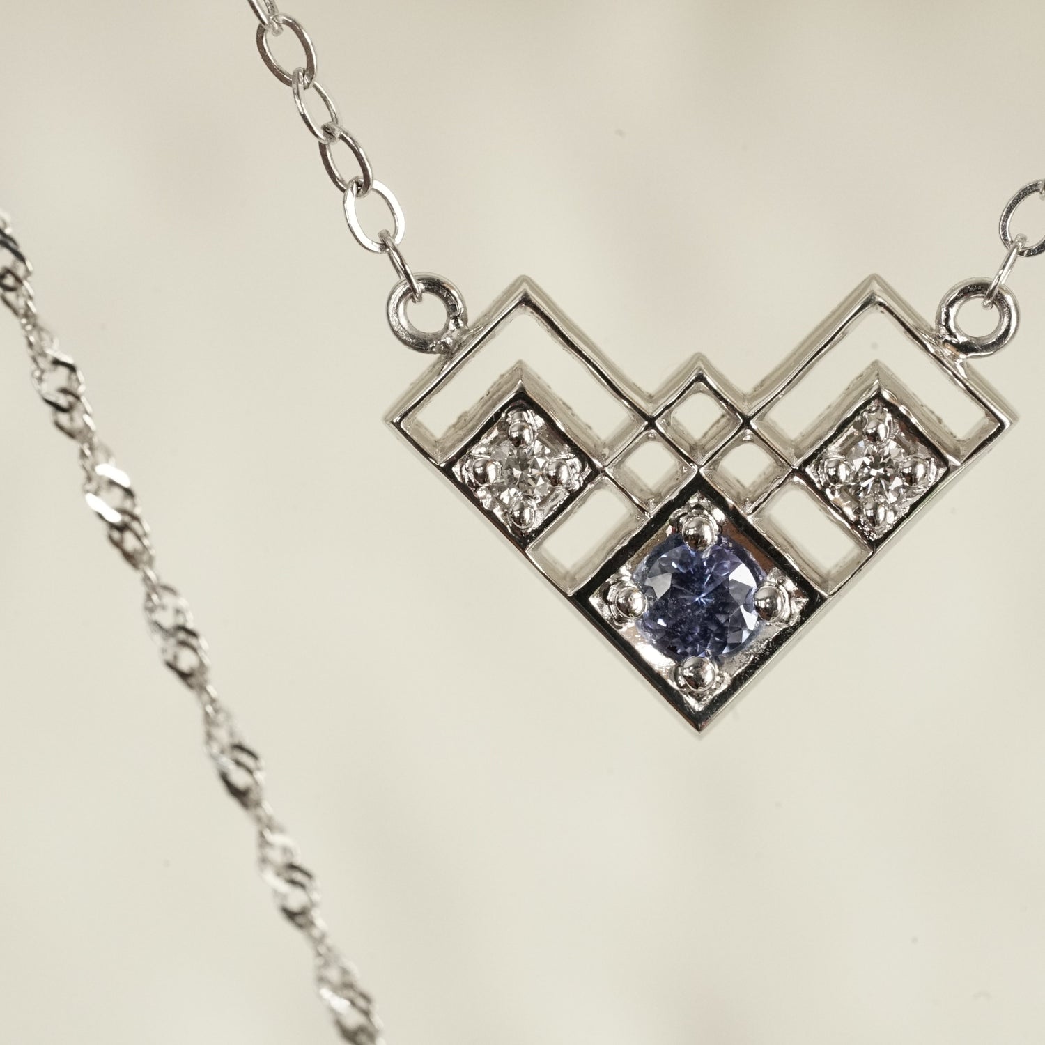 a macro photo of a white gold pendant with a light blue sapphire and 2 white diamonds in a semi heart shape art deco inspired pendant suspended on an open link white gold chain