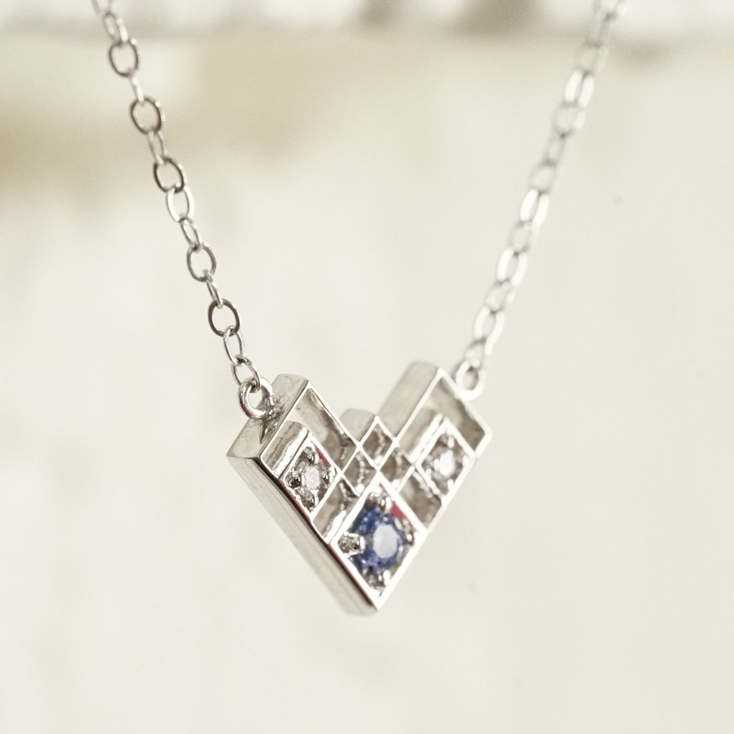 a slight profile view macro photo of a white gold pendant with a light blue sapphire and 2 white diamonds in a semi heart shape art deco inspired pendant suspended on an open link white gold chain