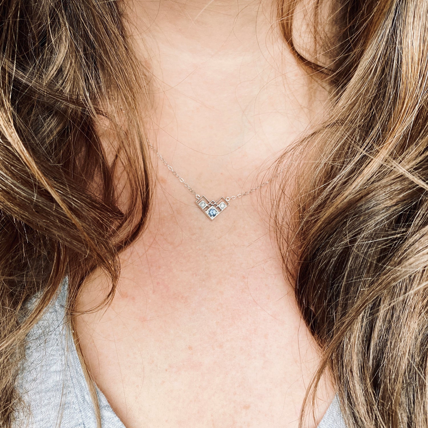 a white gold pendant with a light blue sapphire and 2 white diamonds in a semi heart shape art deco inspired pendant suspended on an open link white gold chain photographed on the neck of a light skinned brunette
