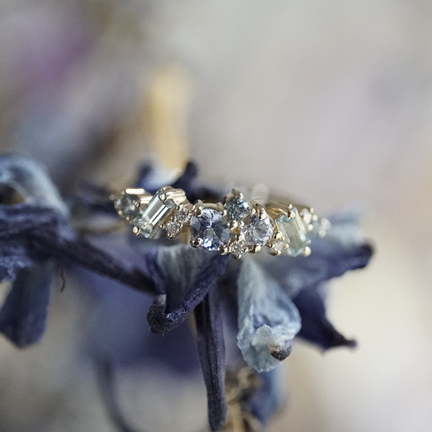 a yellow gold ring with blue gems of various colors sizes and shapes are mixed with round white diamonds and photographed on a dark blue flower