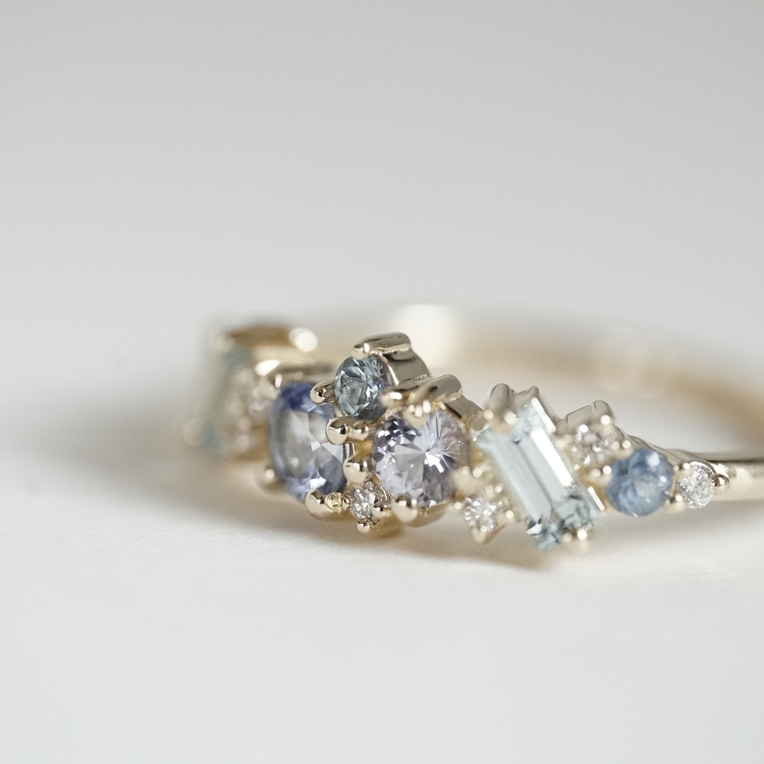a macro photo of the side view of a yellow gold ring with blue gems of various colors sizes and shapes are mixed with round white diamonds on a matte white background