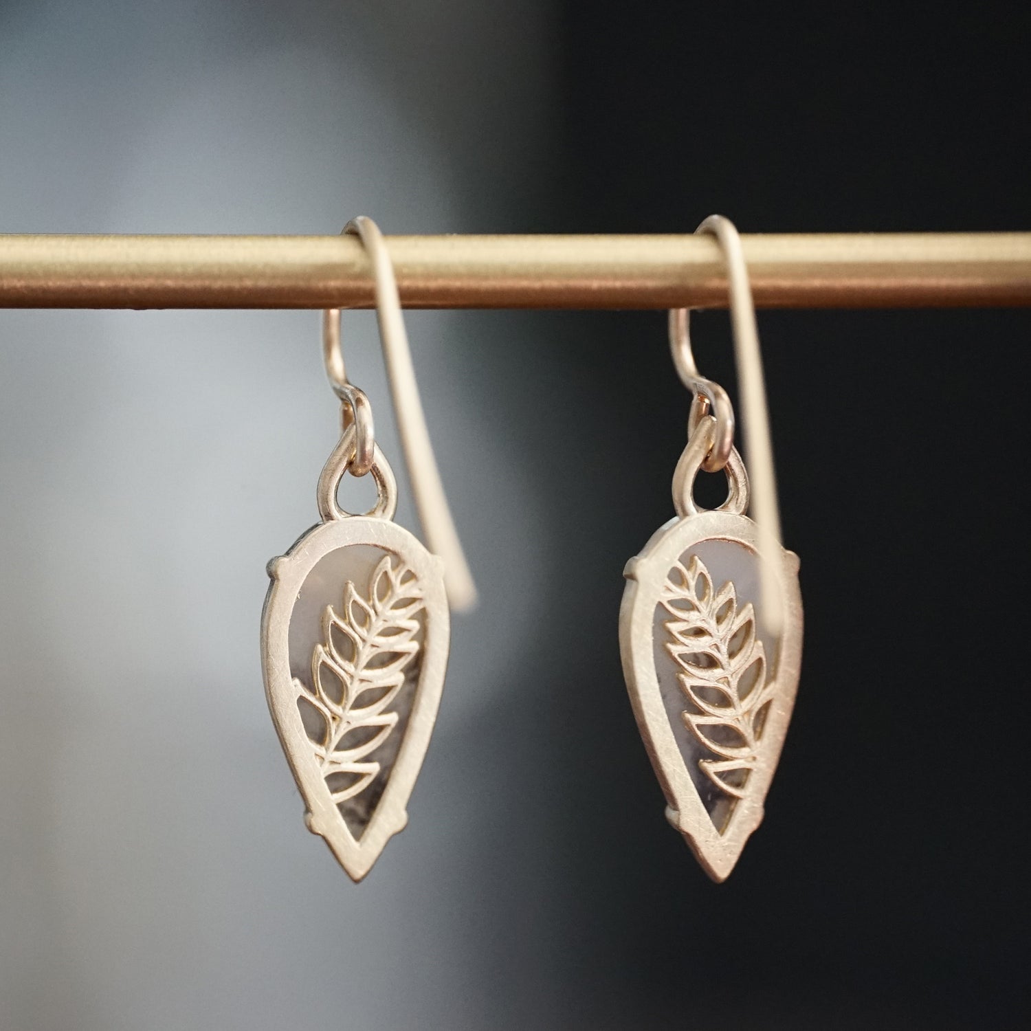 a pair of yellow gold drop earrings with dendritic agates are rotated to show the leaf detailing on the backside an hang on a brass bar