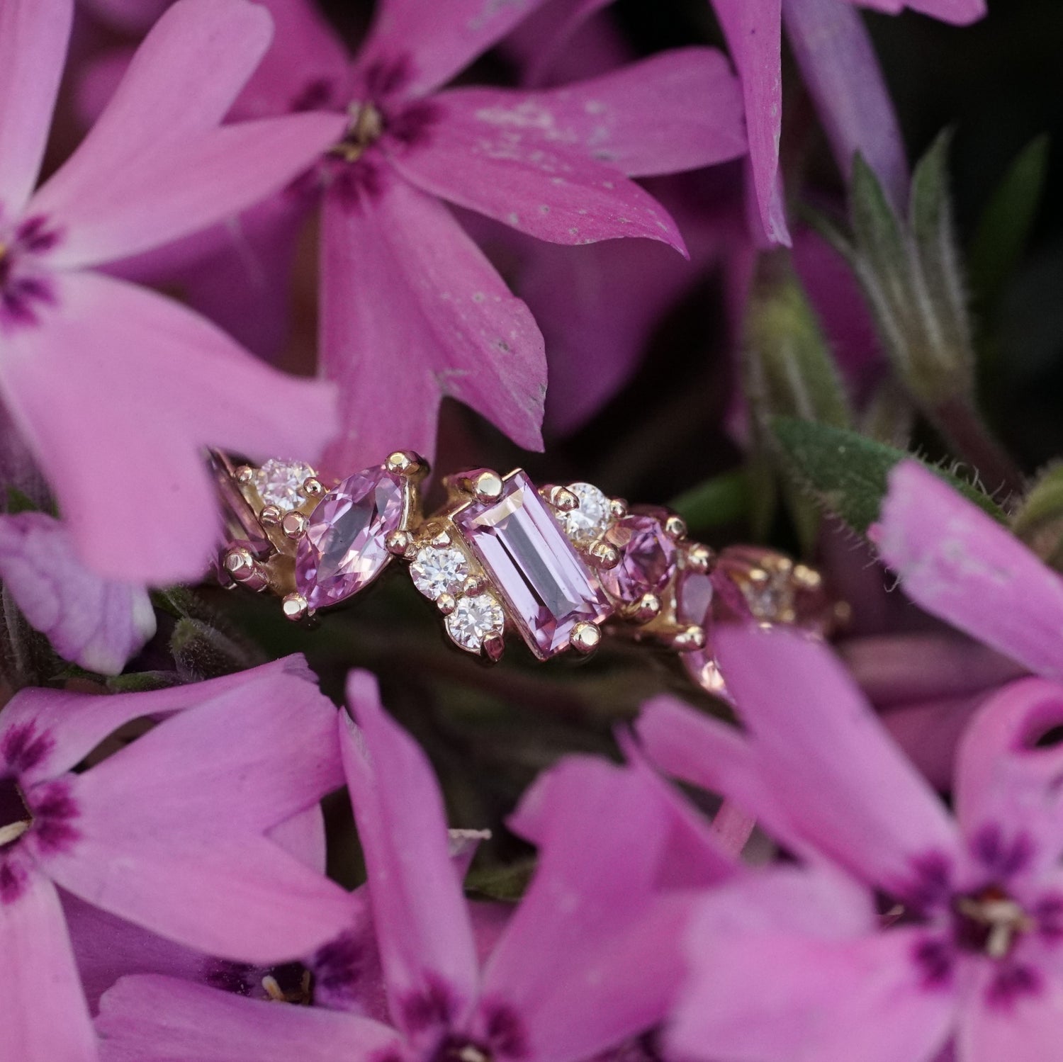 a yellow gold ring with pink sapphires of mixed shapes are paired with round white diamonds in a playful design rests in a field of pink flowers