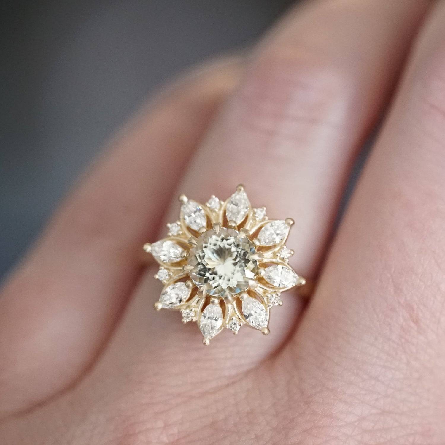 A yellow gold ring with a very light green prasiolite center stone and mixed cut diamond halo on a light skinned left hand