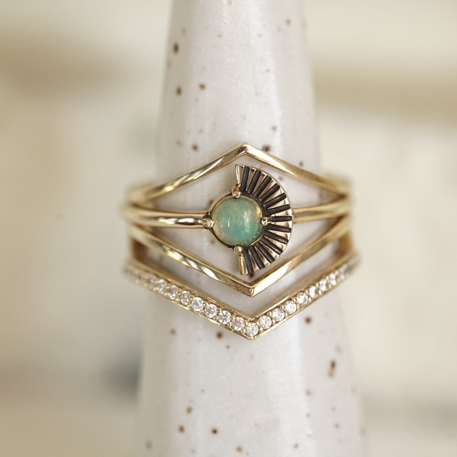a cabochon opal ring with deco fan element to one side is stacked with v shape shadow bands, one with diamonds and 2 without on a white ceramic display