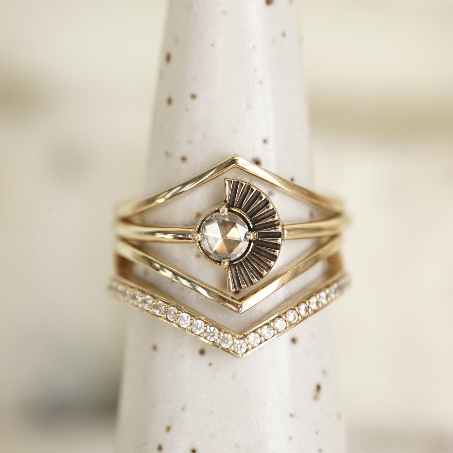 a rose cut white diamond ring with deco fan element to one side is stacked with v shape shadow bands, one with diamonds and 2 without on a white ceramic display