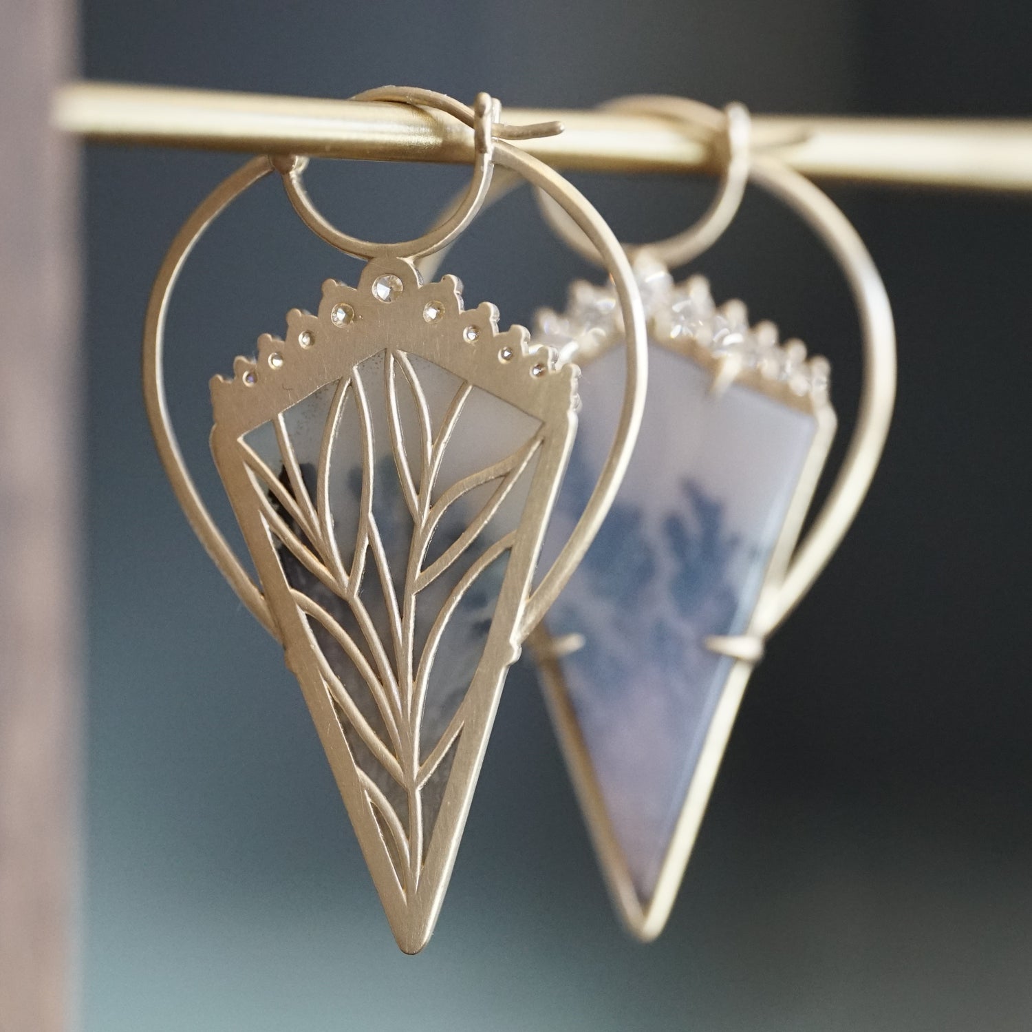 a pair of yellow gold earrings with kite shape dendritic agates and round diamonds hangs on a brass bar one is rotated to show the detail leaf design on the back