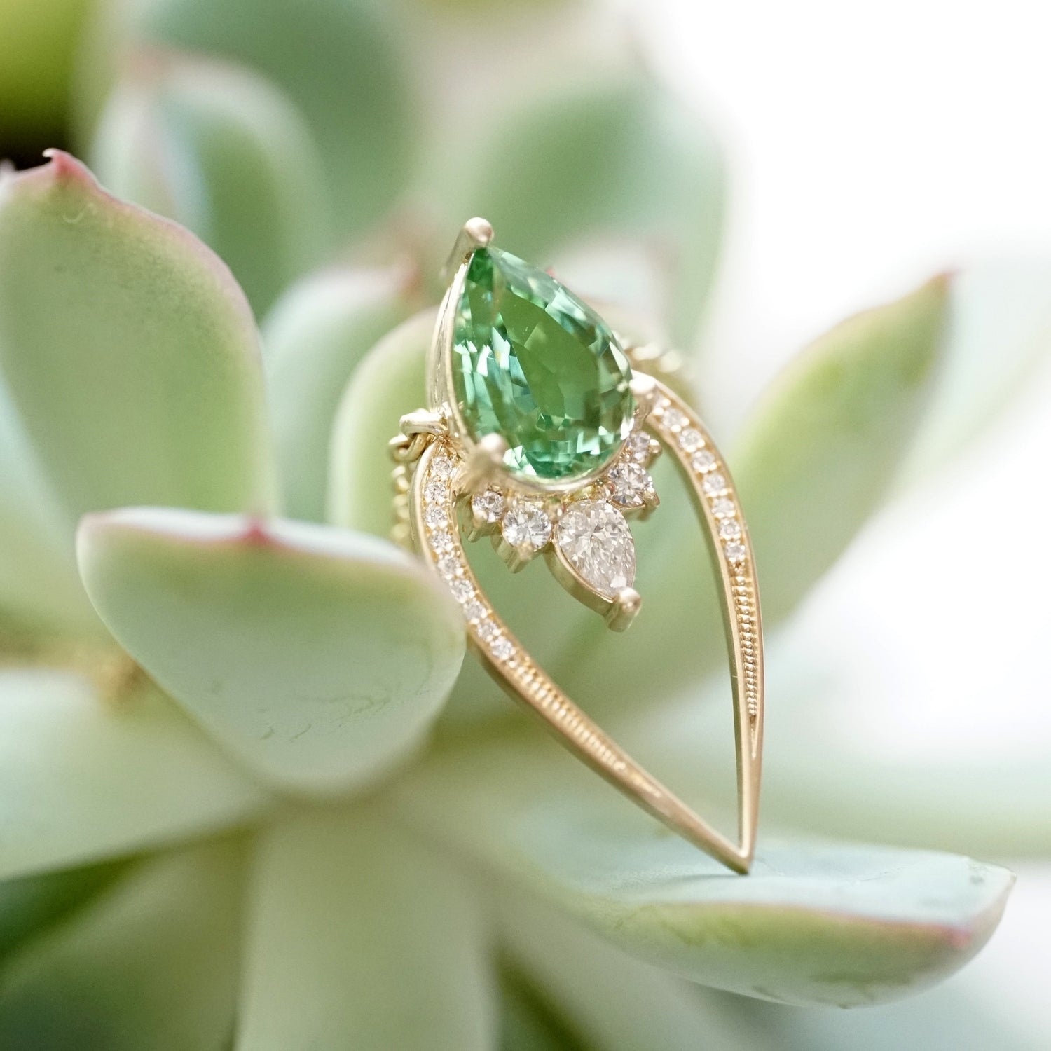 a yellow gold pendant with a bright green tourmaline is accented with vibrant white diamonds and displayed in a succulent plant