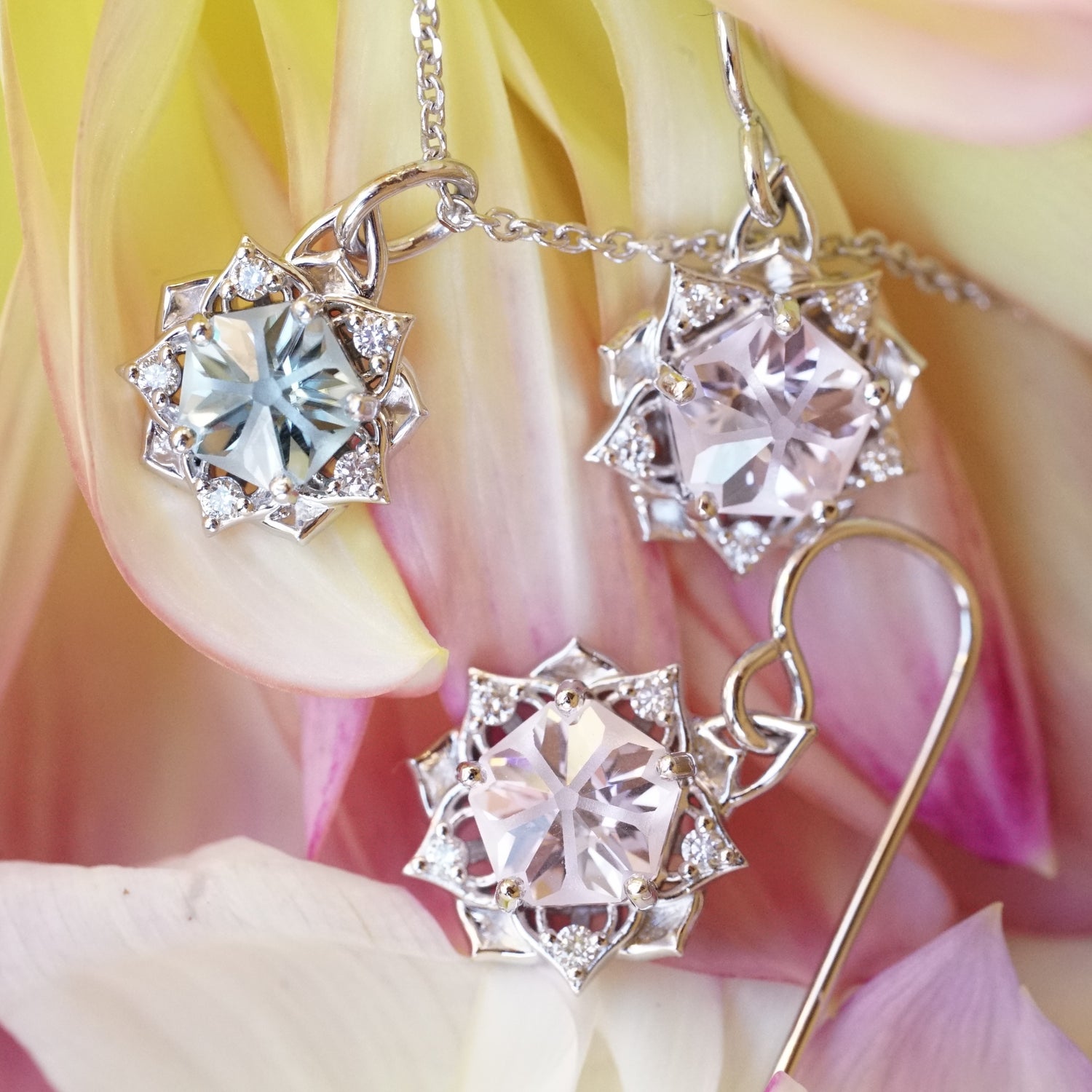 A pair of white gold and pink YAG and diamond flower earrings lay next to a white gold blue zircon diamond flower pendant on pink and yellow flower petals