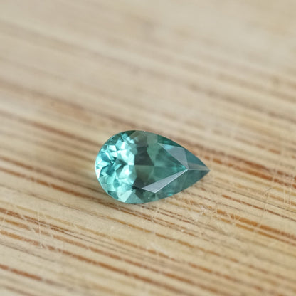 a 0.64 ct vibrant teal tourmaline in a pear shape rests on a wood base