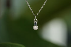 A delicate silver pendant necklace featuring a small clear teardrop-shaped stone and a larger round clear stone, suspended from a fine silver chain against a blurred green background.
