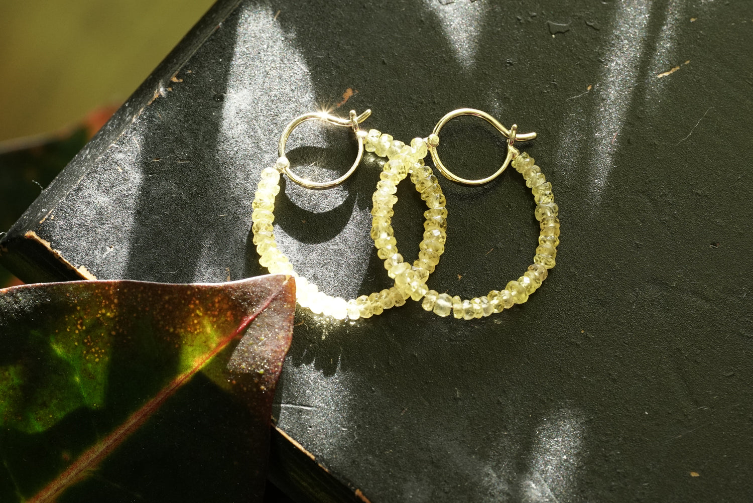 Pair of hoop earrings with faceted yellow sapphire beads on a dark surface with a leaf.