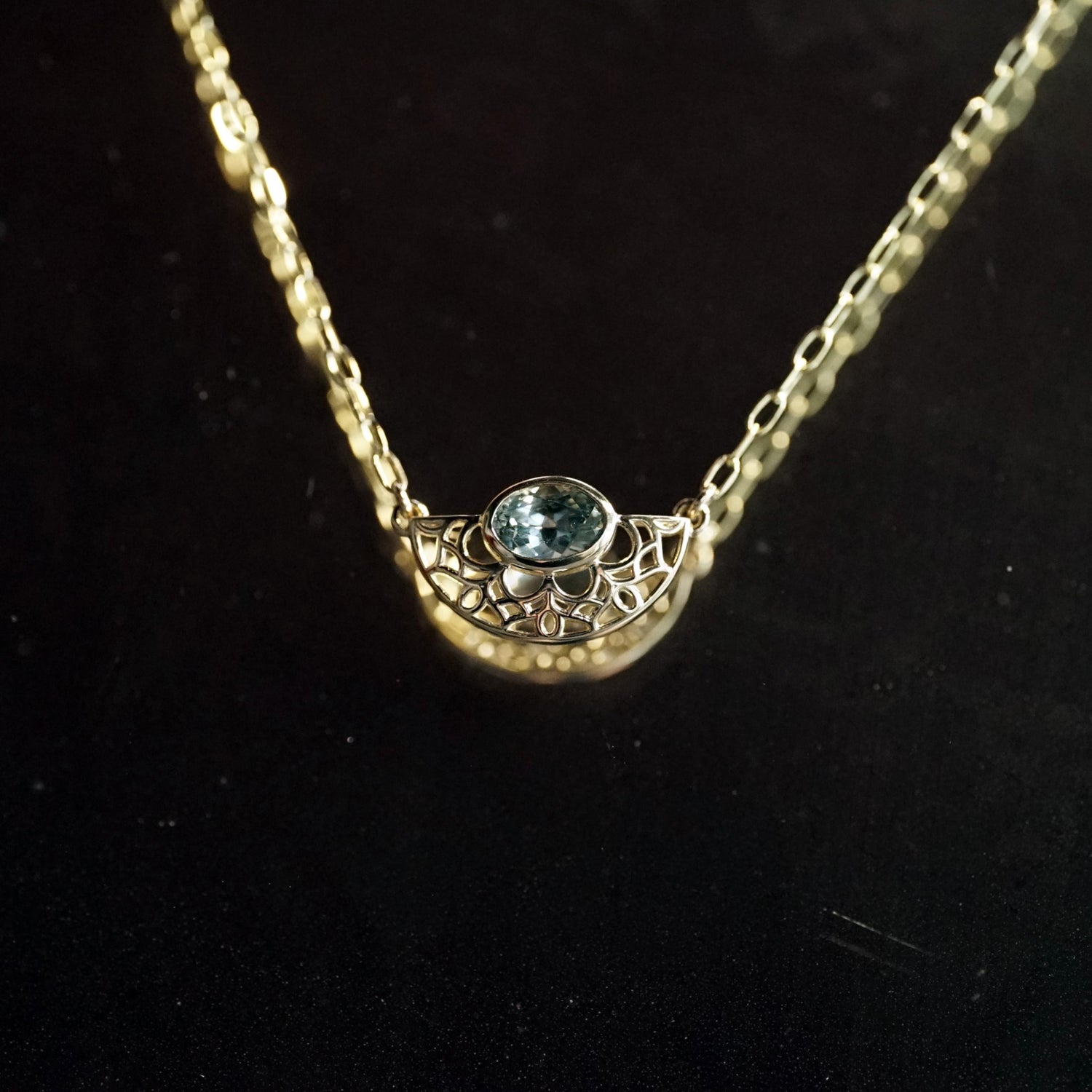 A yellow gold and bezel seat seafoam color sapphire on a light open link chain hanging in front of black mirrored background