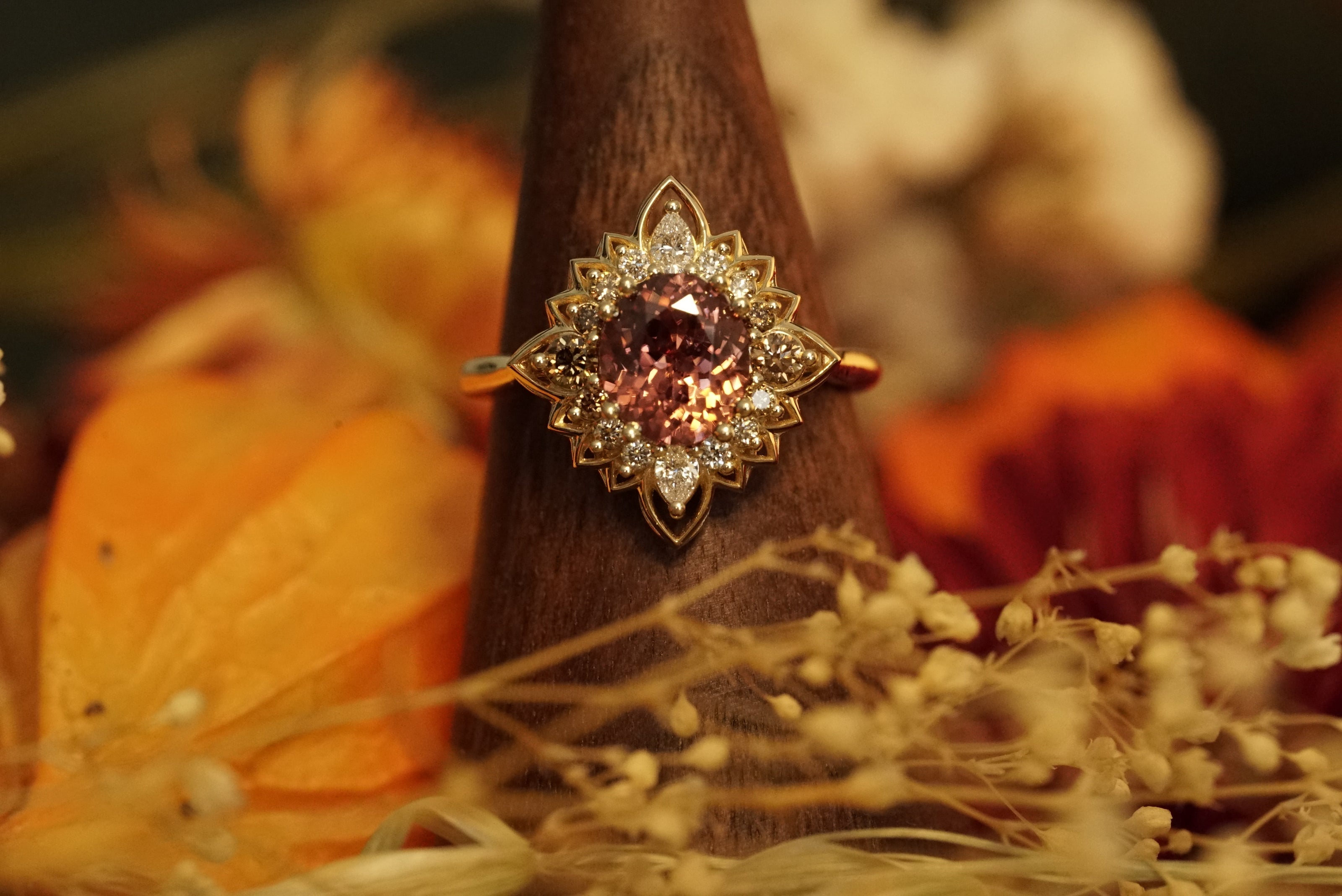 a magnificent 18K yellow gold ring with an oval chocolate zircon center stone surrounded by round and pear shape diamonds which color fade from brown to white in a floral halo shown on a wood display in warm fall foliage