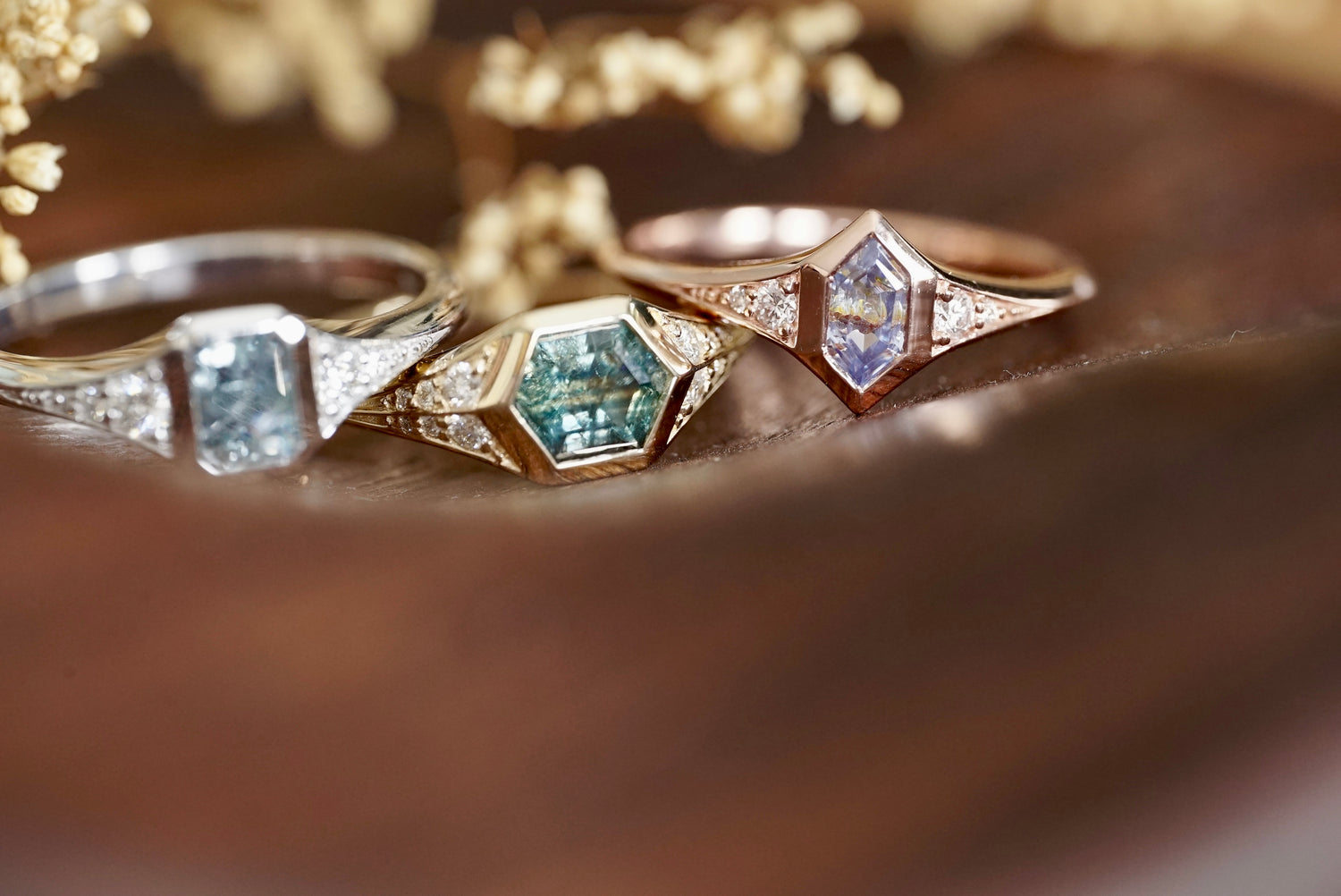 Three sapphire and diamond rings in 14k gold on a wooden surface with flowers in the background