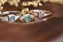 Three sapphire and diamond rings in 14k gold on a wooden surface with flowers in the background