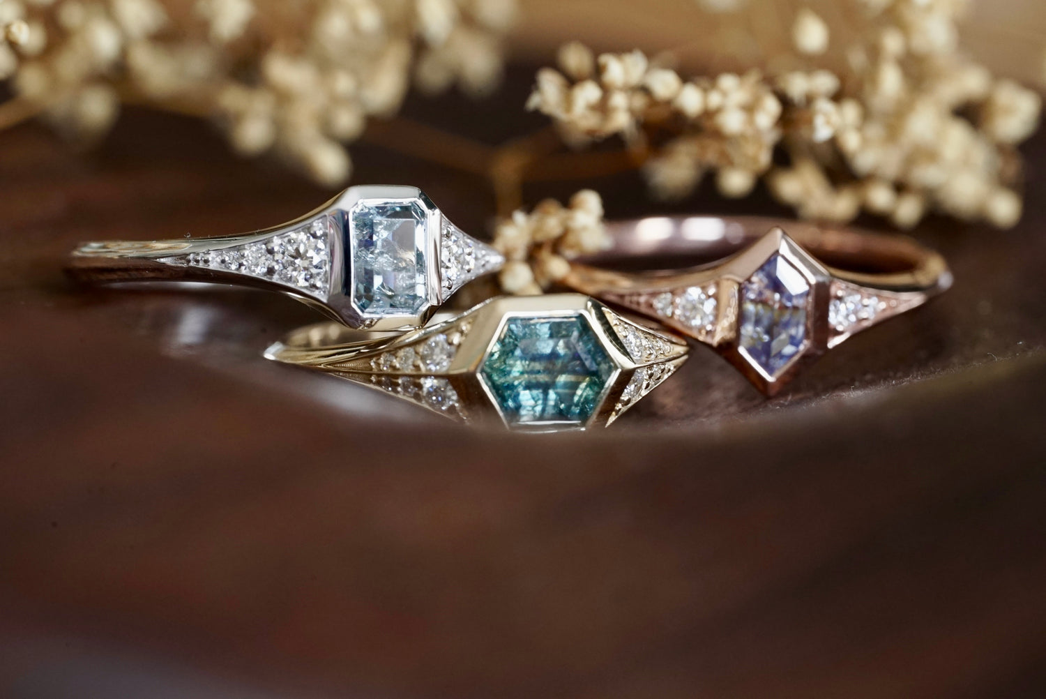 Three sapphire and diamond rings in 14k gold on a wooden surface with flowers in the background