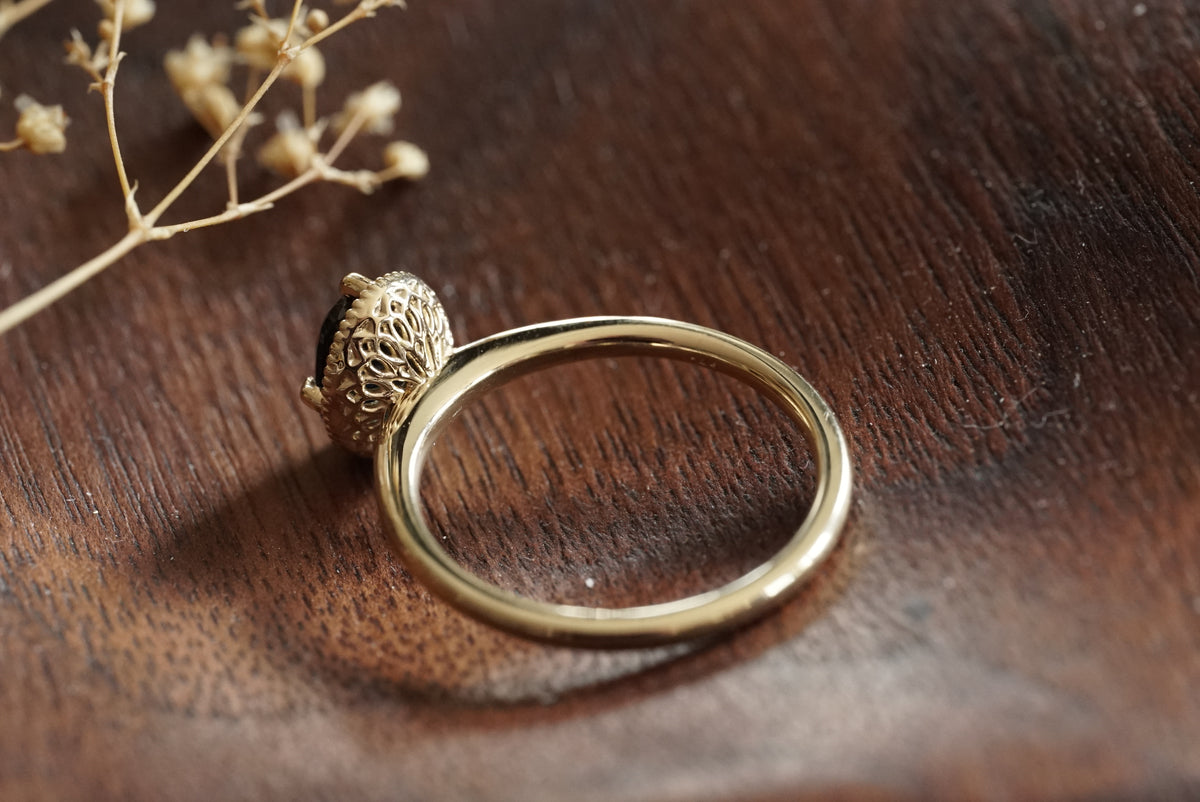 A gold ring with a textured, floral-like filigree basket resting on a dark brown wooden surface with dried flowers in the corner.