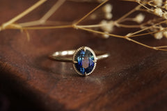 A gold ring with an oval blue gemstone resting on a dark wooden surface with dried flowers in the background.