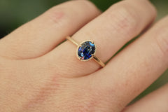 A close-up of a gold ring with a deep blue oval gemstone, worn on a person's finger against a soft, blurred green background.