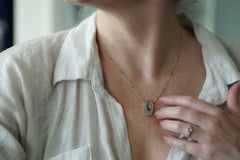 A close-up lifestyle shot of a woman wearing a delicate gold chain necklace with an oval agate pendant, paired with a light-colored linen shirt.