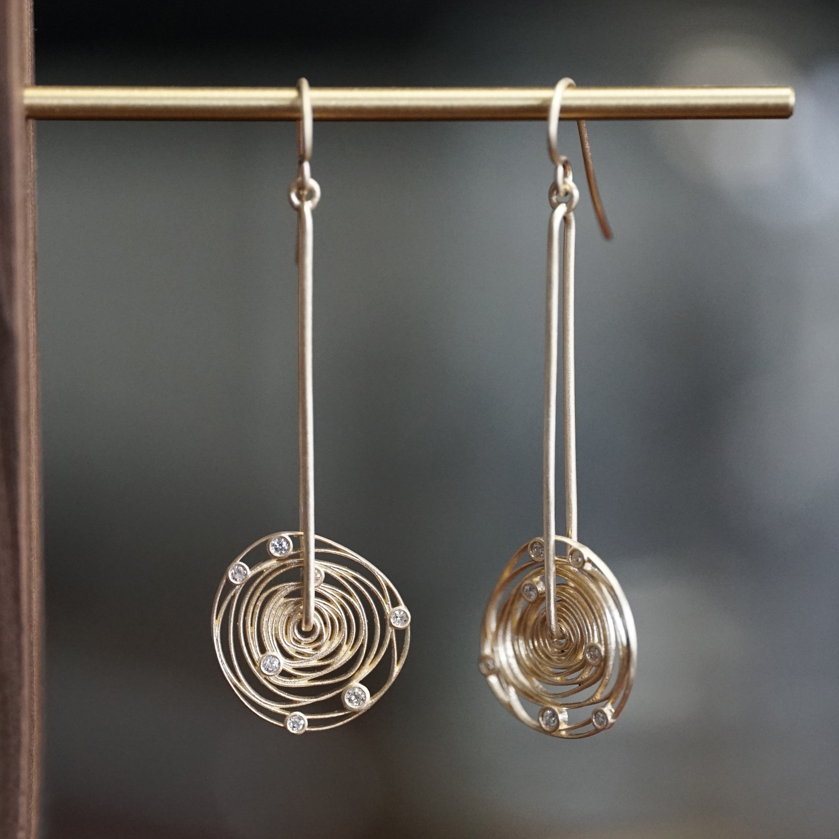 a pair of yellow gold and diamond drop spinning cosmos earrings hangs on a brass bar