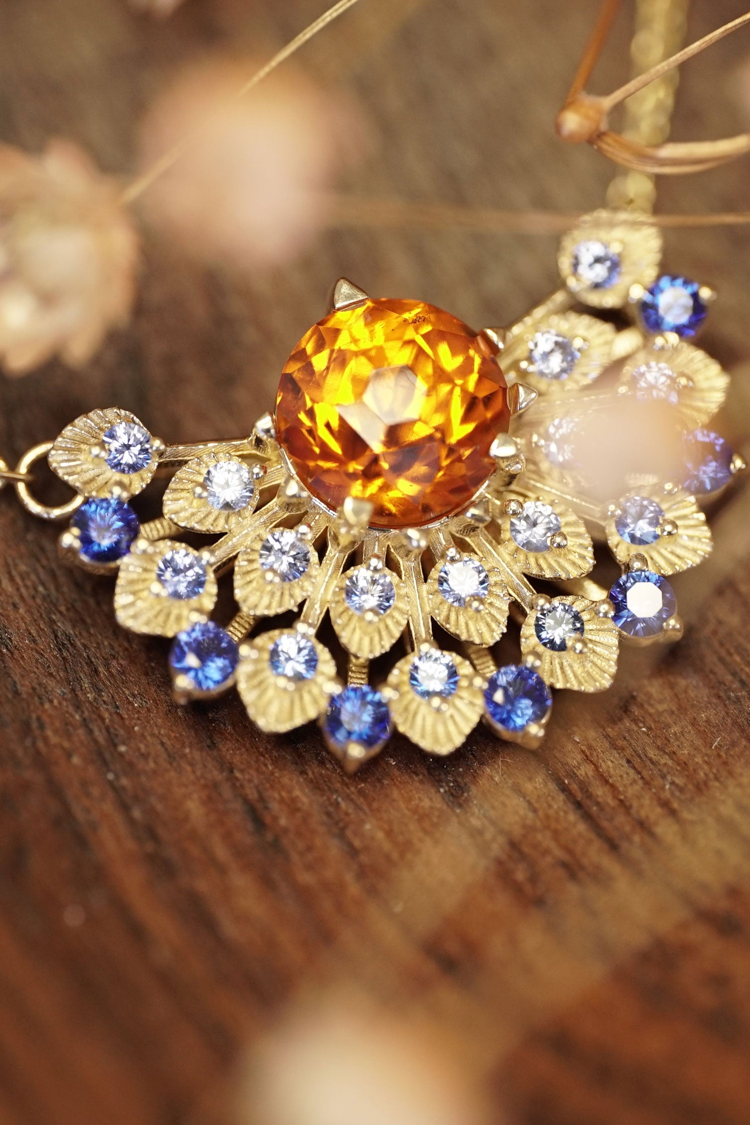 a yellow gold pendant with a vibrant orange garnet center stone and ombre blue sapphires rests on a wood base