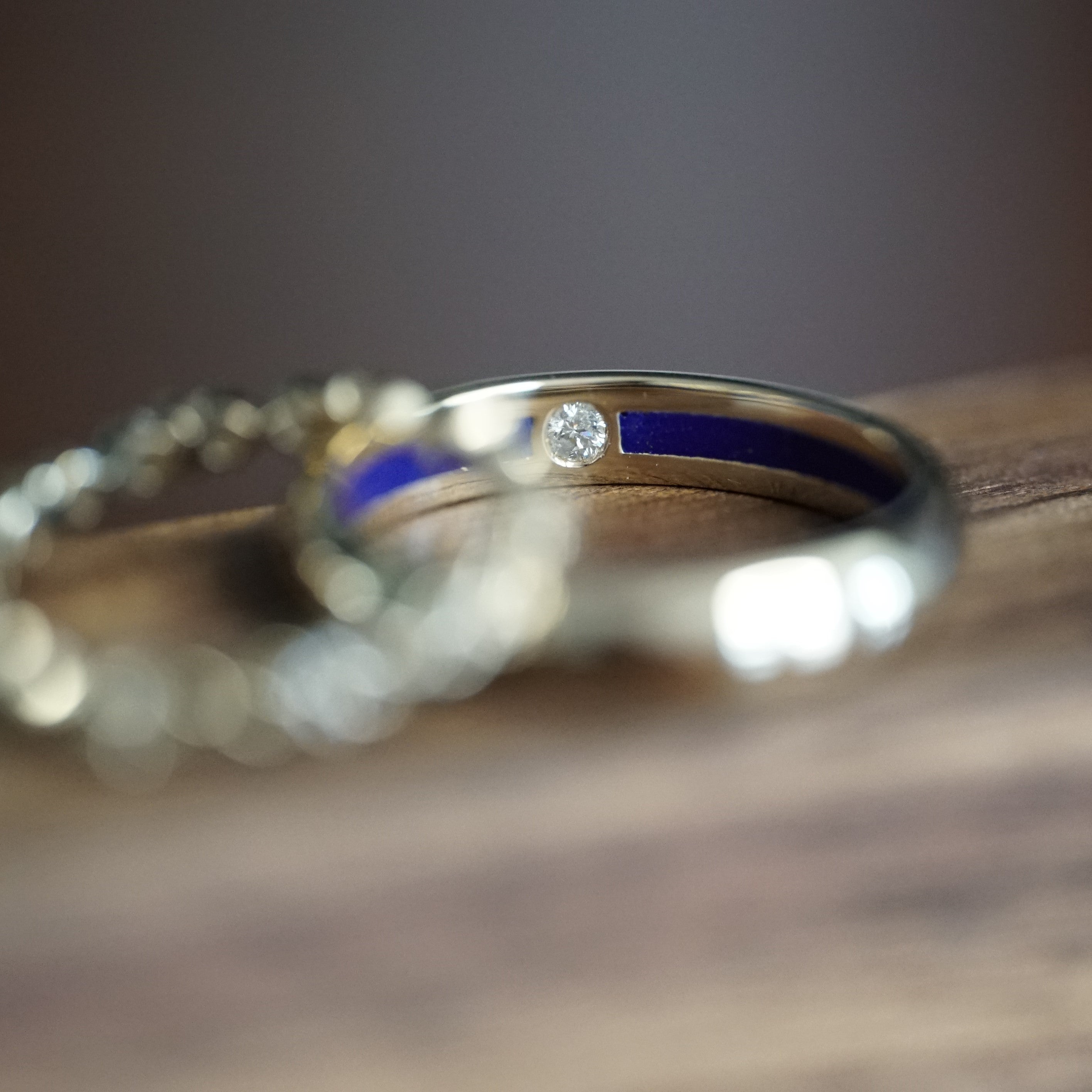 a macro photo of a yellow gold gents wedding band with a diamond and blue resin inside on a wood display