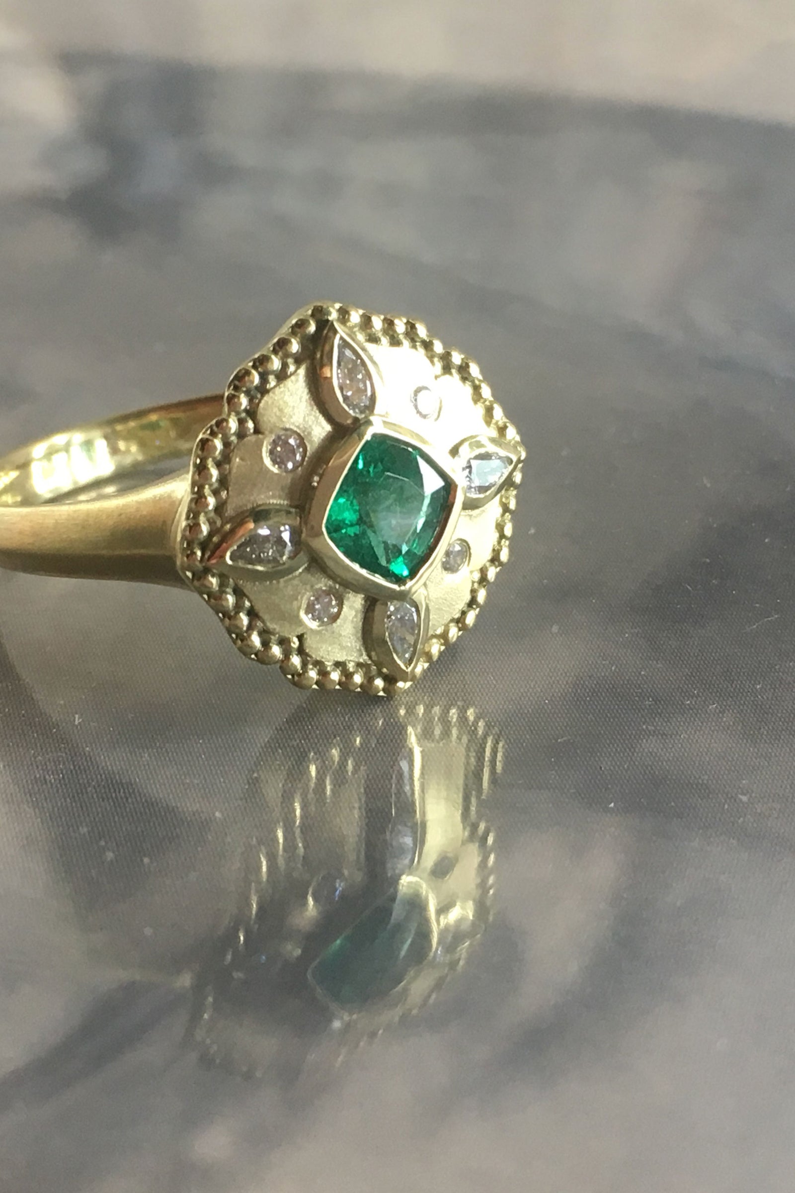 a yellow gold emerald and diamond ring on a grey reflective base