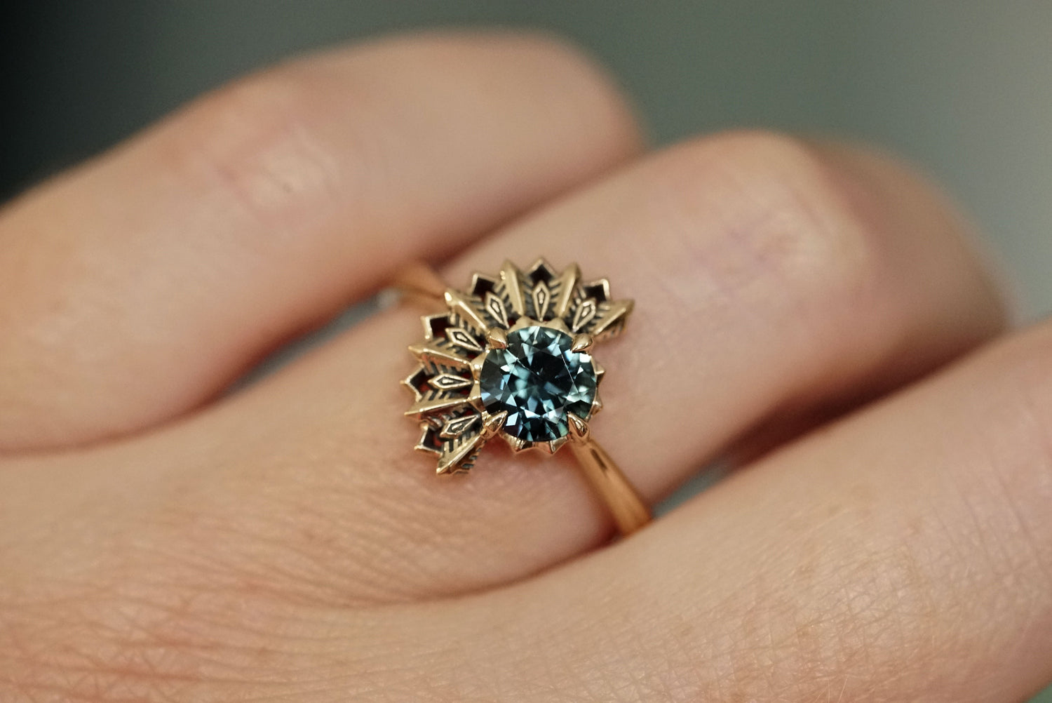 14k yellow gold ring with a deep teal sapphire gemstone on a finger against a neutral background