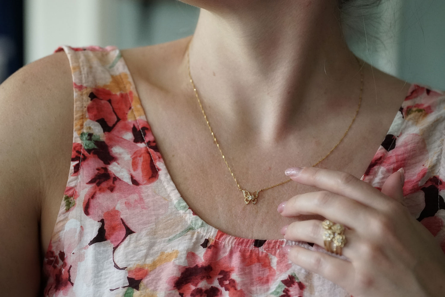 A close-up lifestyle shot of a woman wearing a delicate gold chain necklace with a small bow pendant, paired with a pink and white floral print top.