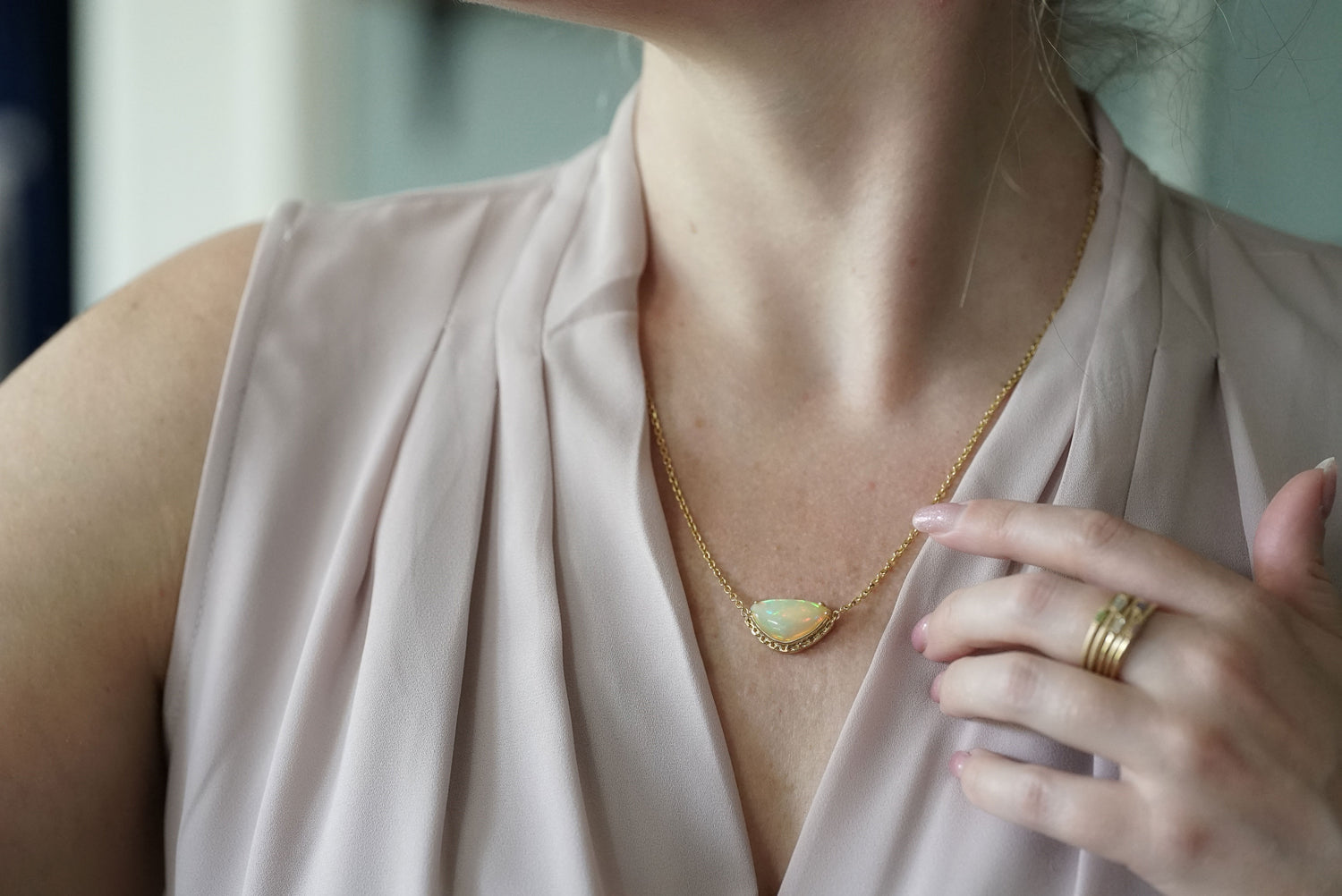 A close-up lifestyle shot of a woman wearing a delicate gold chain necklace with a teardrop-shaped iridescent opal pendant, paired with a light blush pink draped top.