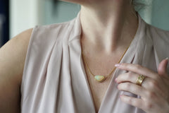 A close-up lifestyle shot of a woman wearing a delicate gold chain necklace with a teardrop-shaped iridescent opal pendant, paired with a light blush pink draped top.