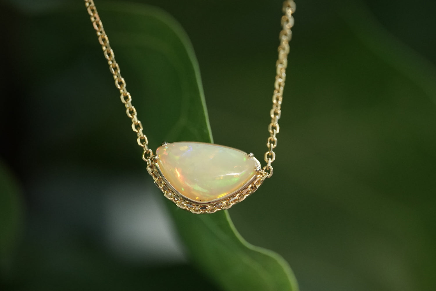 A gold chain necklace with a teardrop-shaped white opal pendant, photographed against a soft-focus green leaf background.