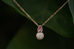 Gold necklace with a teardrop-shaped pendant featuring a pink gemstone and clear center stone on a dark green leaf background.