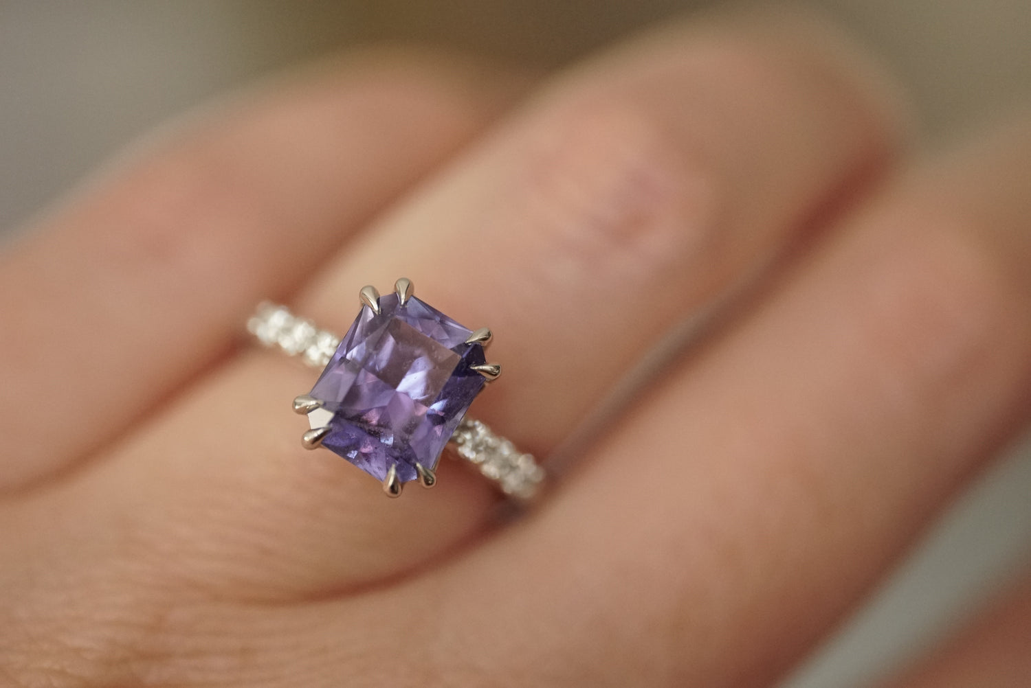 A close-up of a hand wearing a platinum ring with a large rectangular purple sapphire and small diamond accent stones along the band, taken in interior diffused lighting