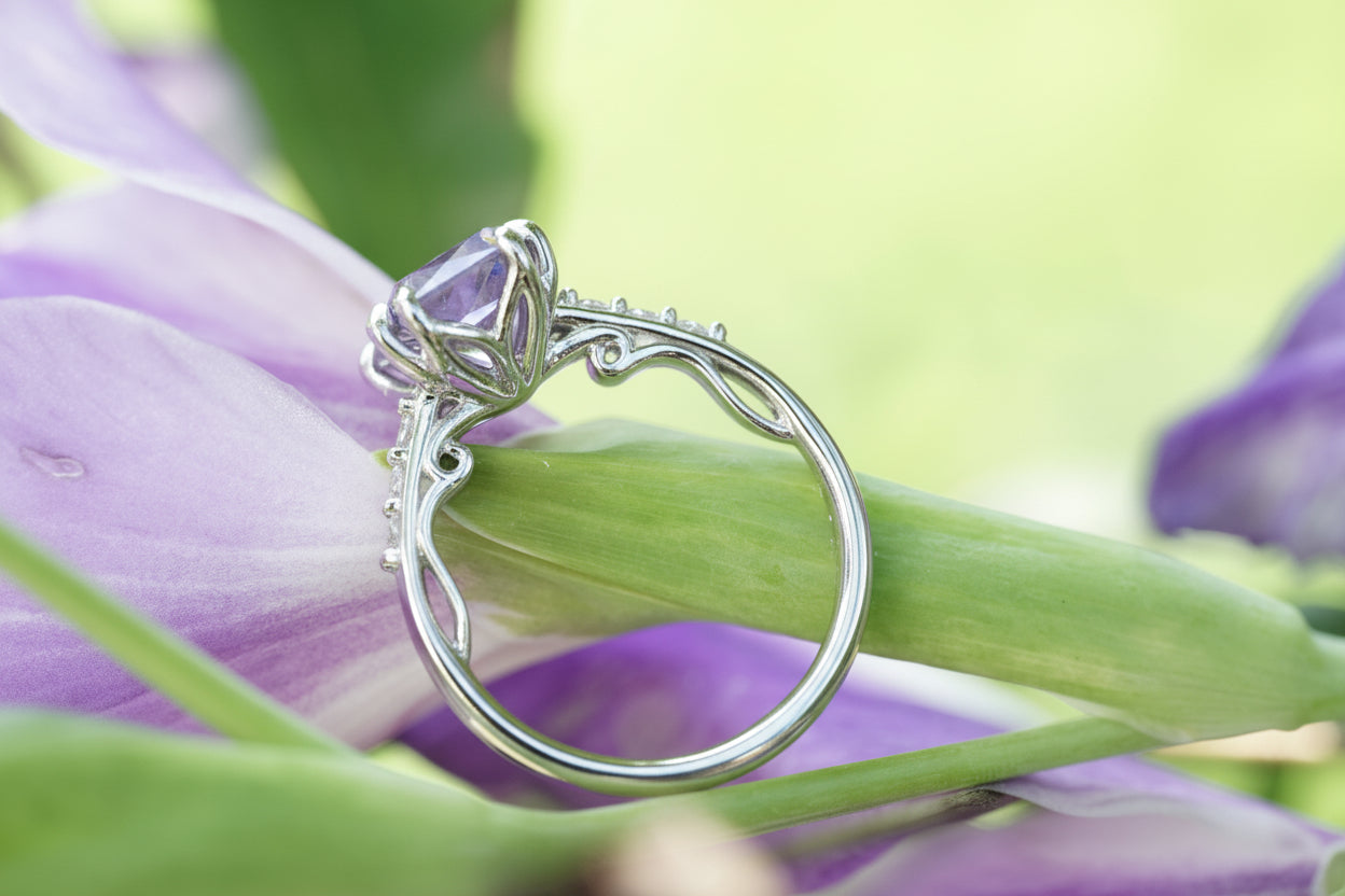 A platinum filigree ring with a purple gemstone, resting on a purple flower with green leaves against a soft-focus green background.