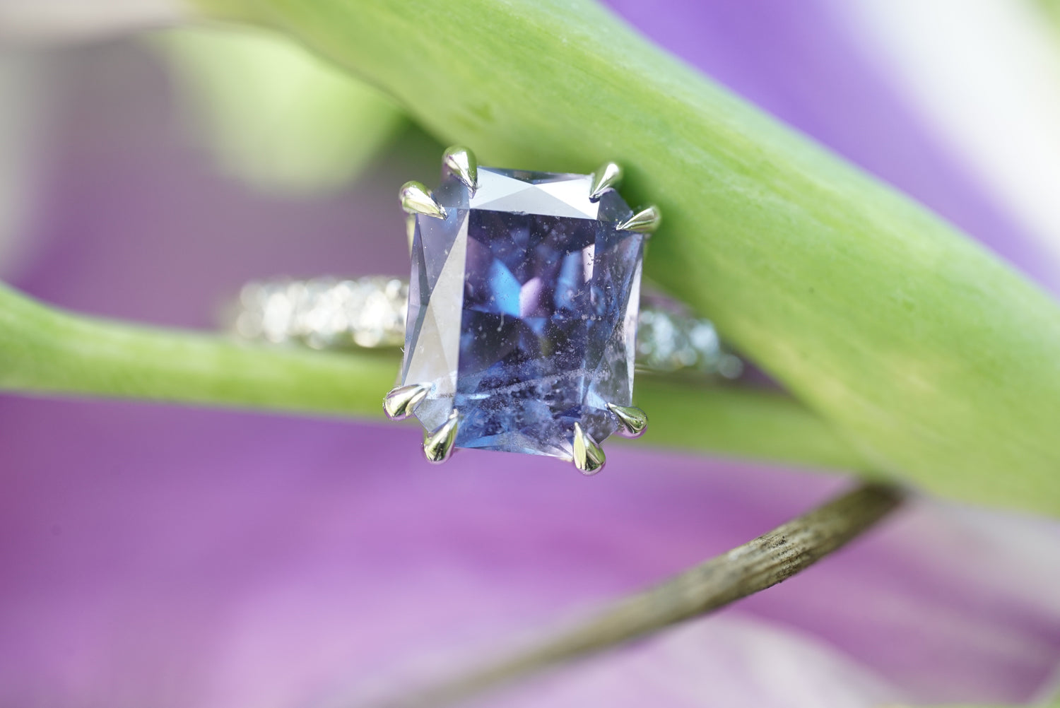 A rectangular purple sapphire ring with a platinum prong setting, shown close-up against a blurred purple and green background.