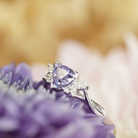 A macro photo of a triangular shaped lavender spinel and diamonds set in a white gold ring laying on a purple flower with a yellow backdrop