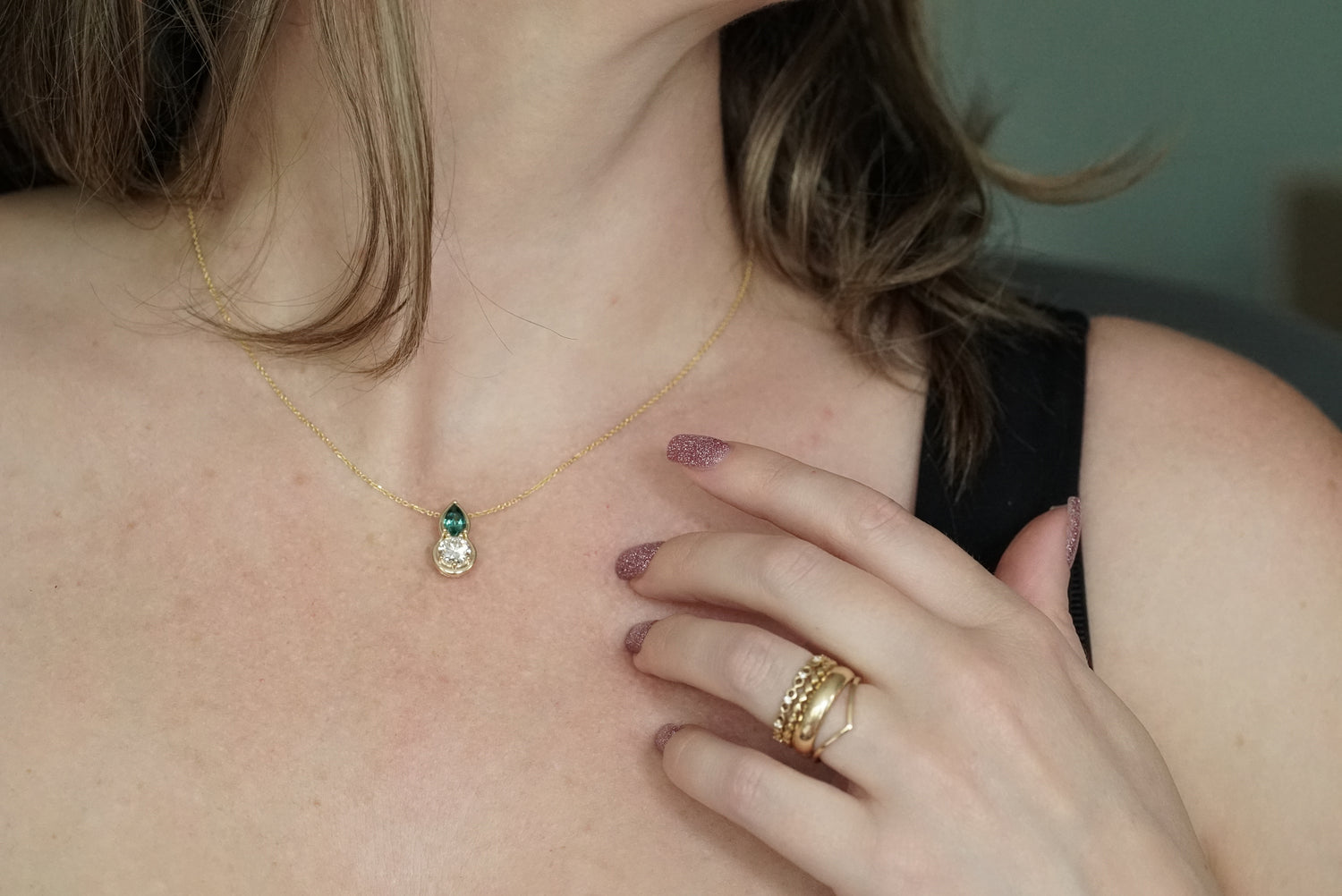 Close-up of a person wearing a gold necklace with a pendant and multiple gold rings on a neutral background.