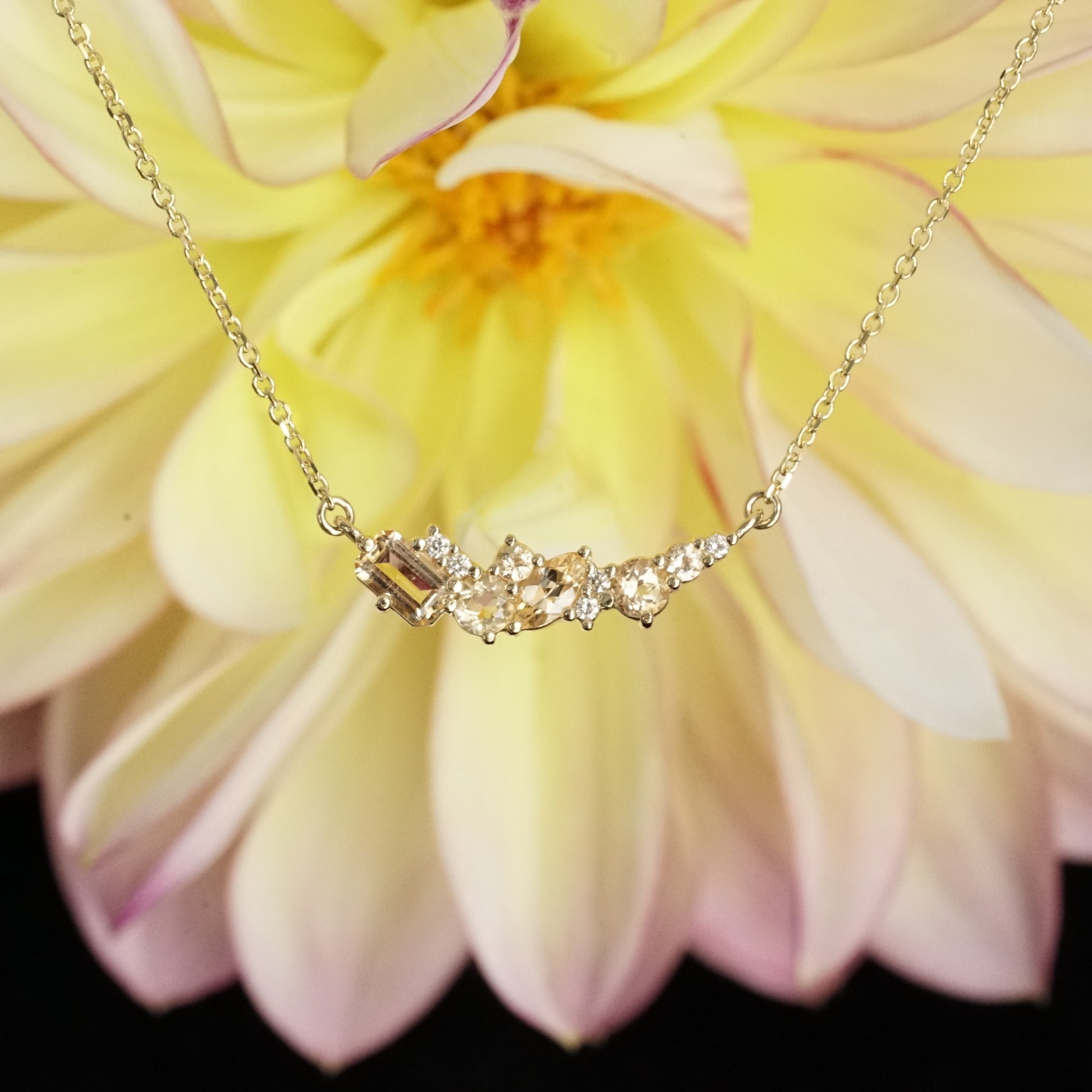 golden topaz of various sizes and shapes are mixed with round white diamonds in a pendant and suspended on an open link yellow gold chain in front of a yellow and pink flower