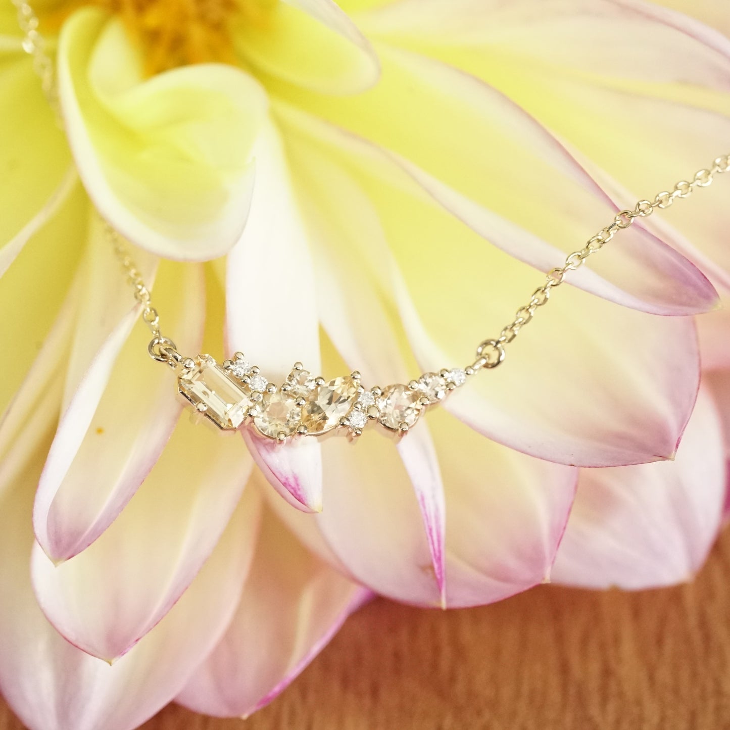 golden topaz of various sizes and shapes are mixed with round white diamonds in a pendant and suspended on an open link yellow gold chain in front of a yellow and pink flower