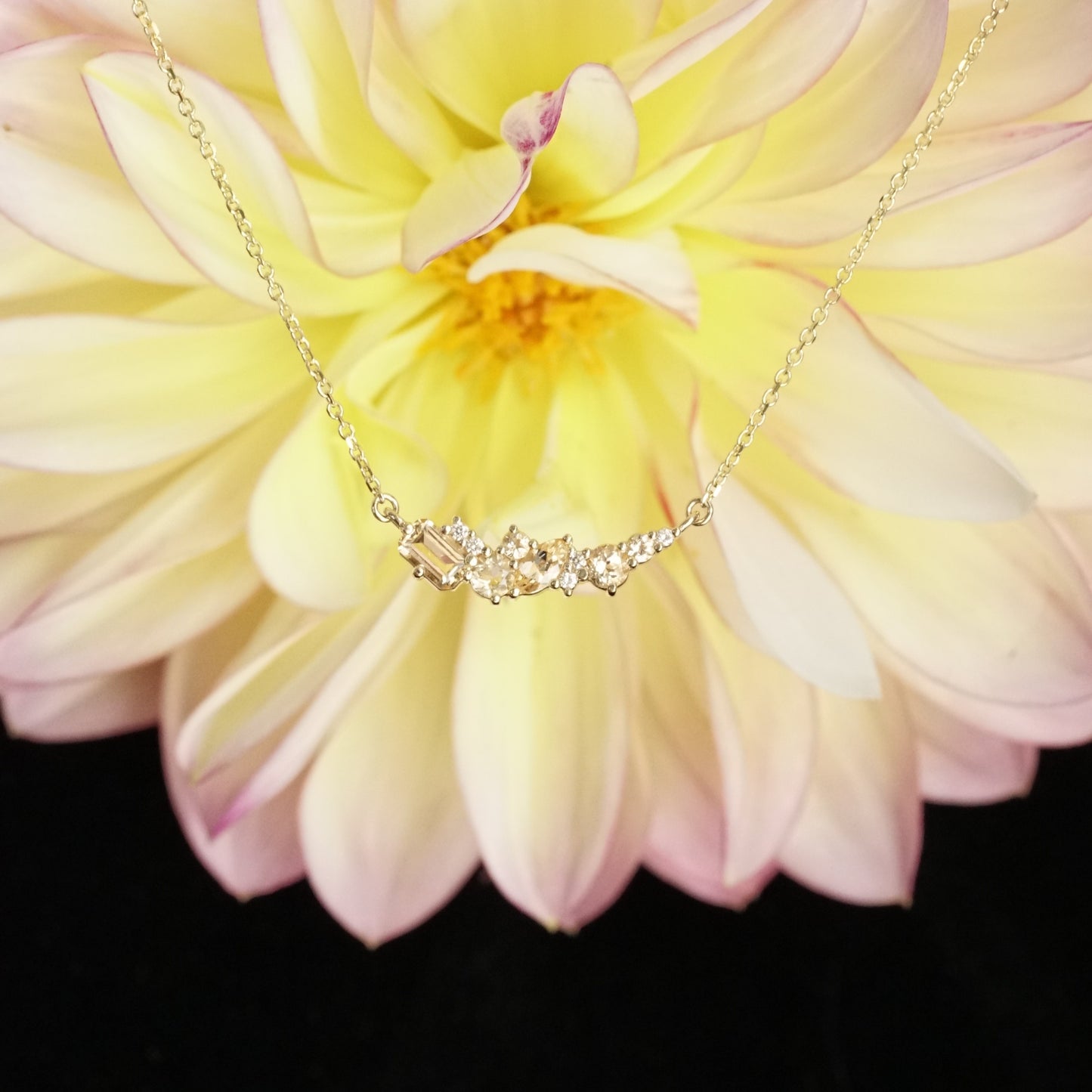 golden topaz of various sizes and shapes are mixed with round white diamonds in a pendant and suspended on an open link yellow gold chain in front of a yellow and pink flower