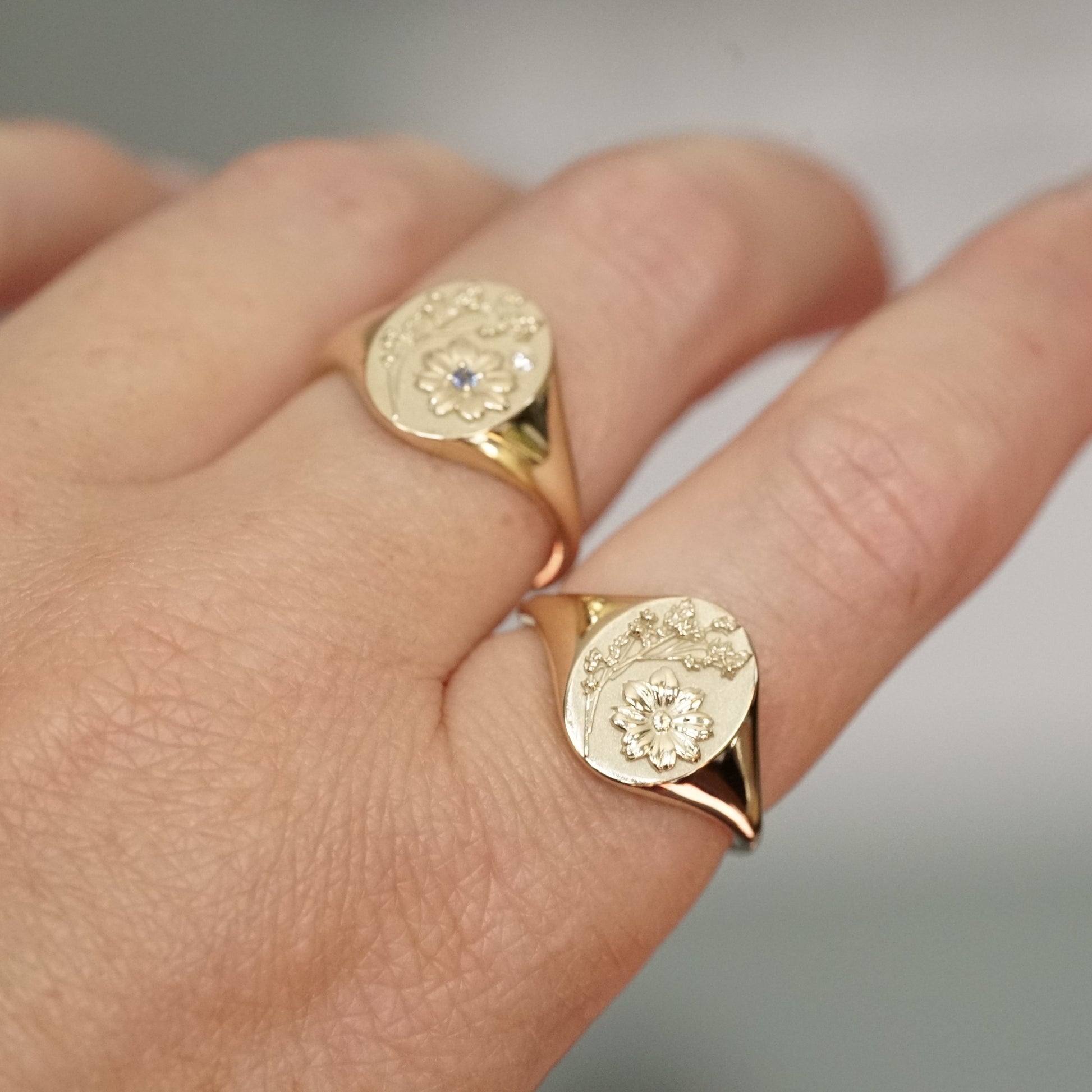 two yellow gold signet rings with floral motifs, one with one light blue sapphire and one diamond accent and the other with no stones, on a light skinned left hand