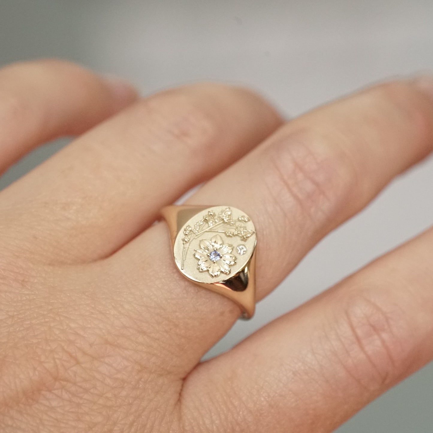 a yellow gold signet ring carved with floral motifs, featuring one light blue sapphire and one diamond accent on a light skinned left hand