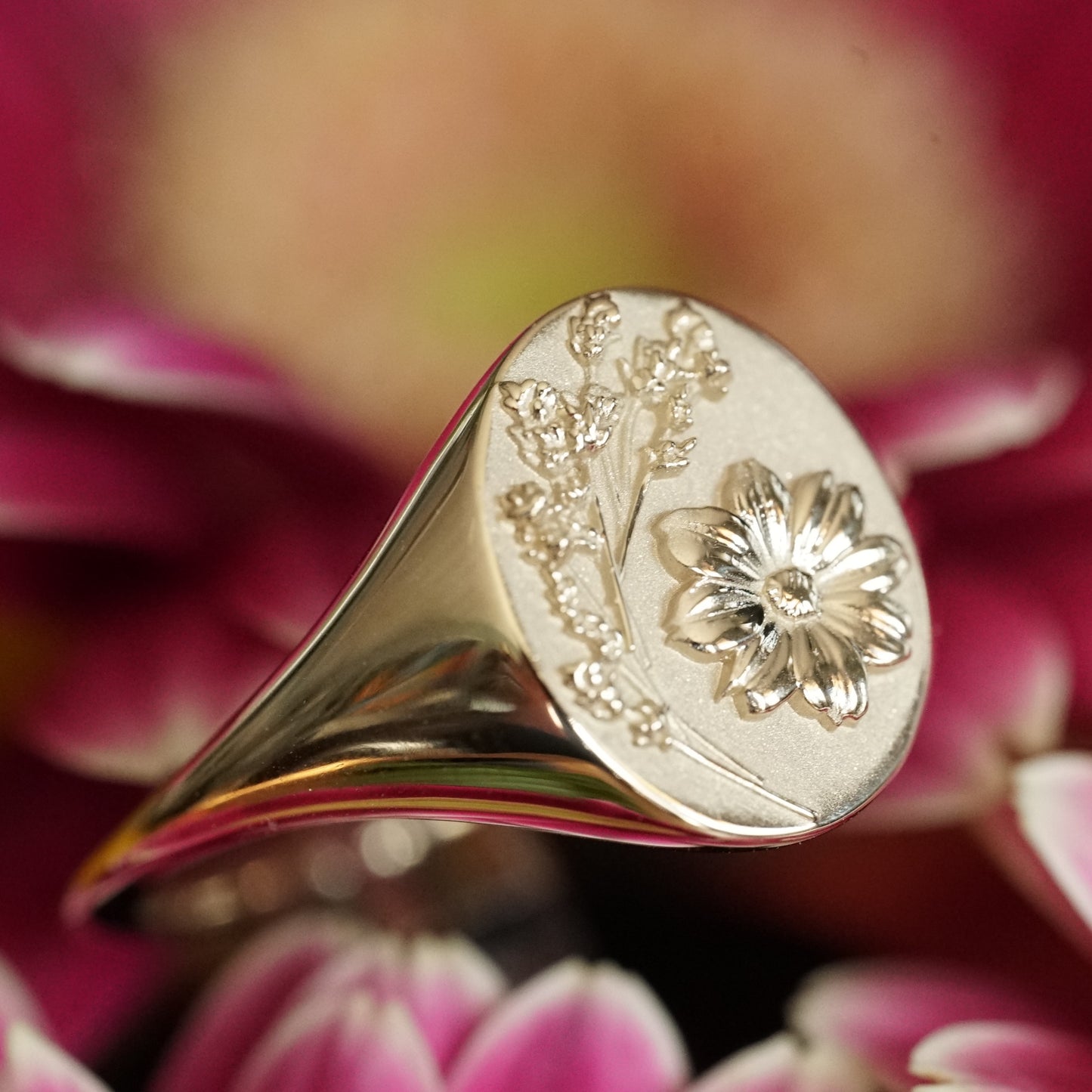 a macro photo of a yellow gold signet rings with floral motifs resting on a pink flower angled in slight profile view