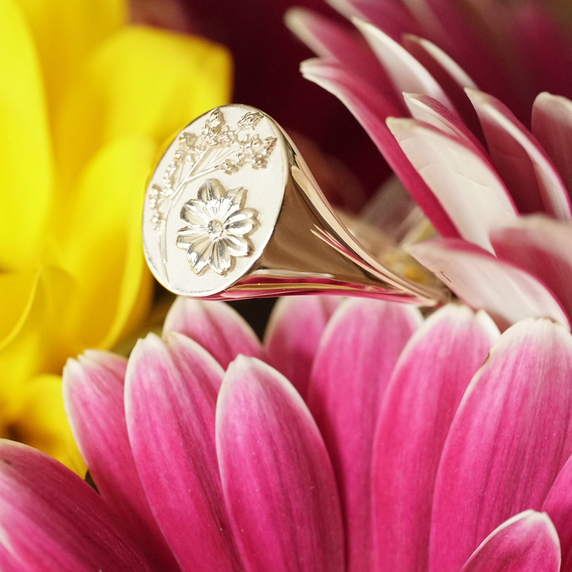 a photo of a yellow gold signet rings with floral motifs resting on a pink flower angled in slight profile view resting in pink and yellow flowers