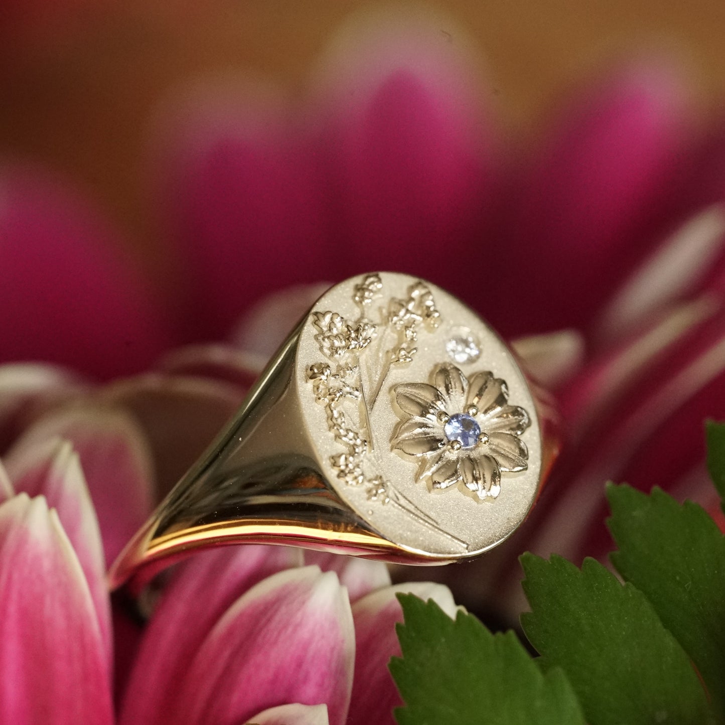 a macro photo of a yellow gold signet rings with floral motifs and accented with one light blue sapphire and one diamond resting on a pink and green flower angled in slight profile view