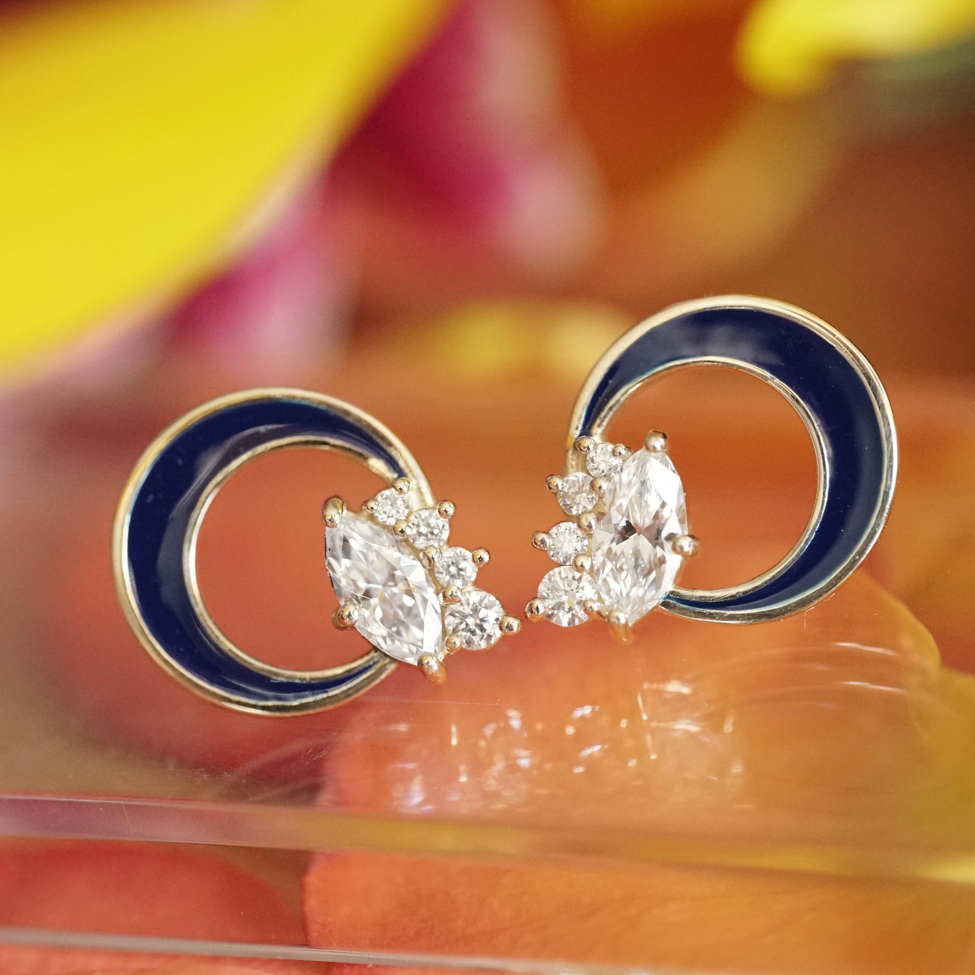 a pair of diamond button earrings with one large marquise and 4 small round diamonds at the base of a crescent shape element filled with blue enamel resting on a reflective base over a mix of orange and yellow flowers